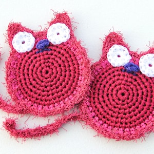 May include: Two handmade, crocheted cat-shaped coasters. The coasters are primarily a deep pink color with white and purple accents for the eyes and nose. Each coaster has a long, fringed tail.