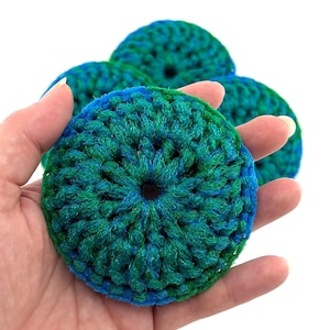 Pode incluir: Uma mão segura vários itens circulares de crochê. Os itens são uma mistura de fios verde-azulado, azul e verde. O fio é bem tecido e cada item tem um buraco no centro. O fundo é branco.