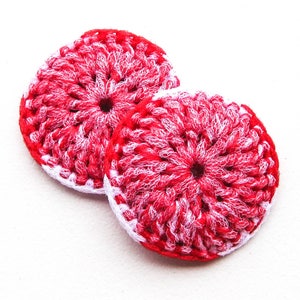 May include: Two handmade, circular crochet items. The items are primarily red with white accents, creating a textured appearance. The edges are finished with a white border. The center of each item has a small hole.