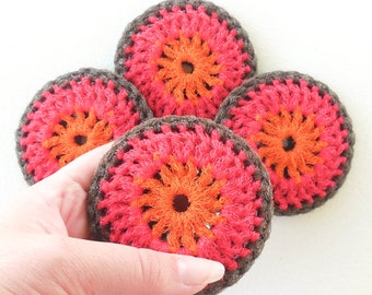 Puede incluir: Cuatro posavasos hechos a mano con ganchillo. Cada posavasos presenta un diseño circular con una combinación de colores vibrantes. El centro es naranja, rodeado por un anillo rojo y un borde marrón oscuro. Los posavasos son sostenidos por una mano.