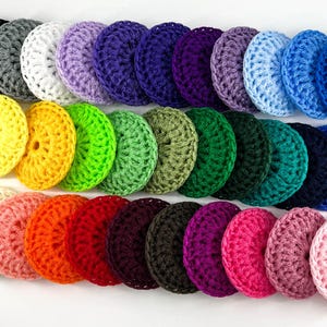 May include: A collection of colorful, crocheted circular items in various shades, including black, gray, white, purple, blue, yellow, green, orange, and pink. Each item has a central hole and a textured surface, suggesting they are dish scrubbers.