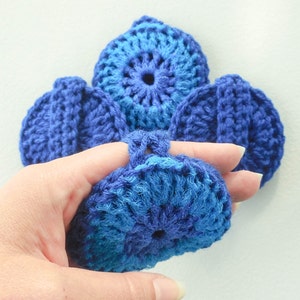 Puede incluir: Primer plano de varios artículos de ganchillo azules. Uno está sostenido por una mano, mostrando un lazo para un dedo. El trabajo de ganchillo presenta un diseño circular con diferentes tonos de azul, creando una apariencia texturizada. Los otros artículos están en el fondo.