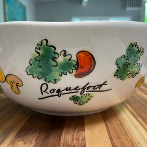 May include: A white ceramic bowl featuring hand-painted illustrations of green leafy vegetables, orange peppers, and yellow mushrooms. The word Roquefort is written in black cursive script on the side of the bowl, which has a smooth, rounded rim.