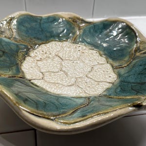 May include: A ceramic decorative plate with a floral design. The plate features a central white flower surrounded by teal-colored leaves. The edges are a light beige color, and the plate has a wavy, organic shape.