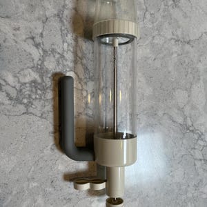 以下が含まれることがあります： 透明な円筒形の容器、ベージュ色のプラスチック製キャップ、トリガー式ノズル付きのベージュ色のベース、グレーのハンドルを備えたキッチン用生地ディスペンサー。細い金属製のロッドが透明なチャンバーの中心を垂直に貫通しています。