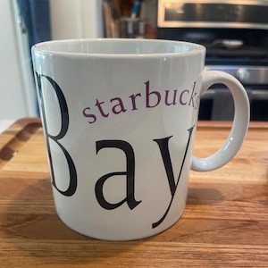 May include: White ceramic coffee mug with the words "starbucks Bay" printed on the side in black and purple lettering. The mug has a curved handle and is sitting on a wooden surface.