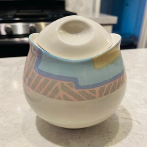 May include: A ceramic sugar bowl with a lid. The bowl is egg-shaped with a white base and a band of pastel colors, including pink, blue, and yellow. The lid is white with a small handle. The bowl has a retro design.