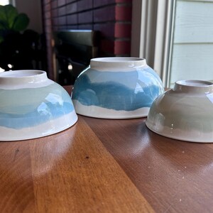 May include: Three ceramic bowls with a hand-painted design. The bowls are in shades of blue, green, and white, with a wave-like pattern. The bowls are sitting on a wooden table.