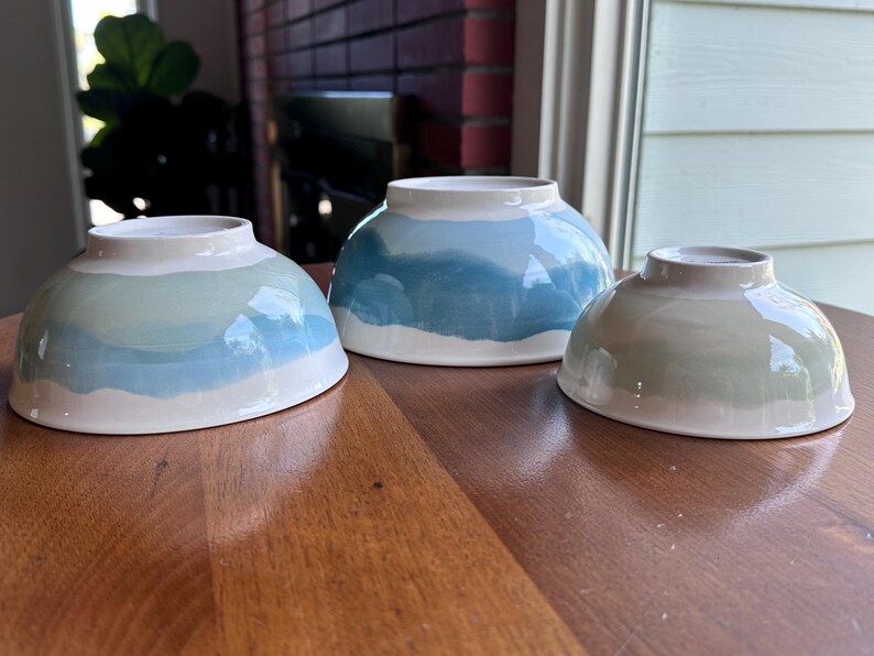 May include: Three ceramic bowls with a hand-painted design. The bowls are in shades of blue, green, and white, with a wave-like pattern. The bowls are sitting on a wooden table.