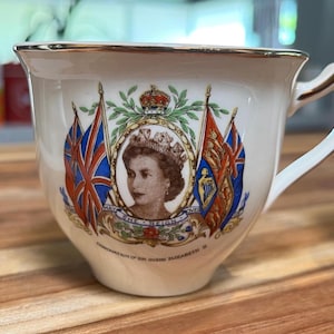 Op de afbeelding: Een crèmekleurige keramische theekop met een goudkleurige rand en handvat. De voorzijde toont een portret van Koningin Elizabeth II, omringd door een kroon, laurierbladeren en twee vlaggen. De tekst onderaan luidt "MAY SHE REIGN" en "CORONATION OF HM QUEEN ELIZABETH II".