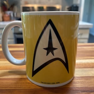 May include: Yellow ceramic mug with a white handle. The mug features the Star Trek insignia in black and white. The bottom of the mug has the text "TM & © 2021 CBS, ARR".