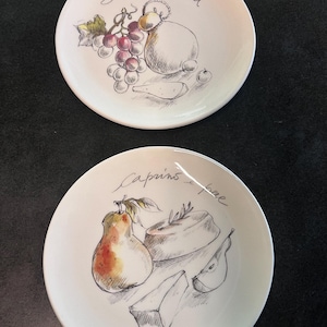 Może przedstawiać: Dwa białe ceramiczne talerze z ręcznie rysowanymi ilustracjami jedzenia. Jeden talerz przedstawia winogrona, ser i tekst "Scamorza e uva". Drugi talerz przedstawia gruszkę, ser i tekst "Caprino e pere".