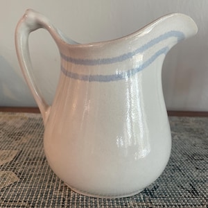 May include: A white ceramic jug with a curved spout and handle. Two light blue stripes encircle the jug's body. The jug has a glossy finish and sits on a patterned surface. This jug is ideal for serving beverages.