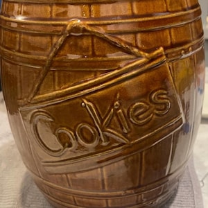 Puede incluir: Bote de galletas de cerámica marrón con forma de barril, con un letrero que dice "Cookies". El bote tiene un acabado brillante y un detalle de cuerda. El diseño del barril presenta líneas horizontales y verticales.