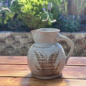 Peut inclure: Une cruche en céramique gris clair avec un corps arrondi et une anse unique. La surface présente une texture nervurée et un motif botanique peint en brun sur le devant. La cruche a un bord large avec un petit bec verseur sur le dessus.