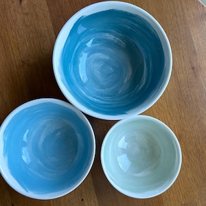 May include: Three ceramic bowls with a white rim and a painted interior. The largest bowl is a deep teal, the medium bowl is a lighter blue, and the smallest bowl is a pale green. The bowls are arranged on a wooden surface.