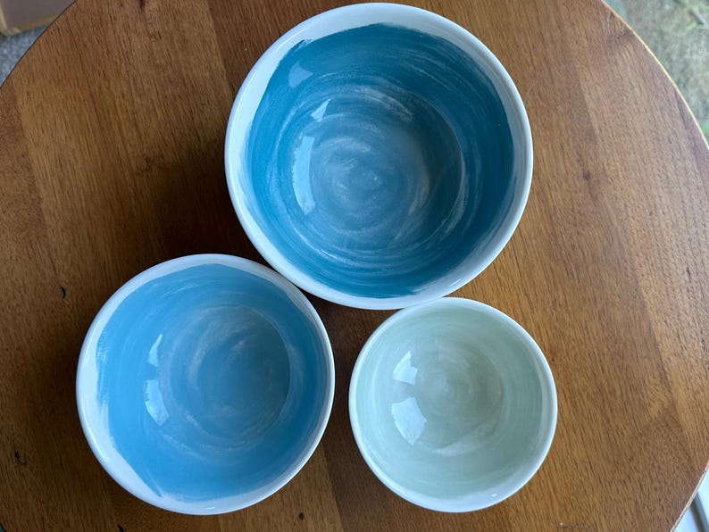 May include: Three ceramic bowls with a white rim and a painted interior. The largest bowl is a deep teal, the medium bowl is a lighter blue, and the smallest bowl is a pale green. The bowls are arranged on a wooden surface.