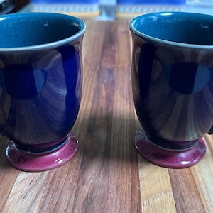 May include: Two ceramic mugs with a dark blue exterior and a teal interior. The base of each mug is a deep red-purple color. The mugs have a curved handle and sit on a wooden surface.