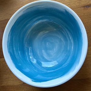 May include: A round ceramic bowl with a white rim and a blue interior. The blue paint has a brushstroke texture, creating a swirling pattern. The bowl is set against a wooden surface.