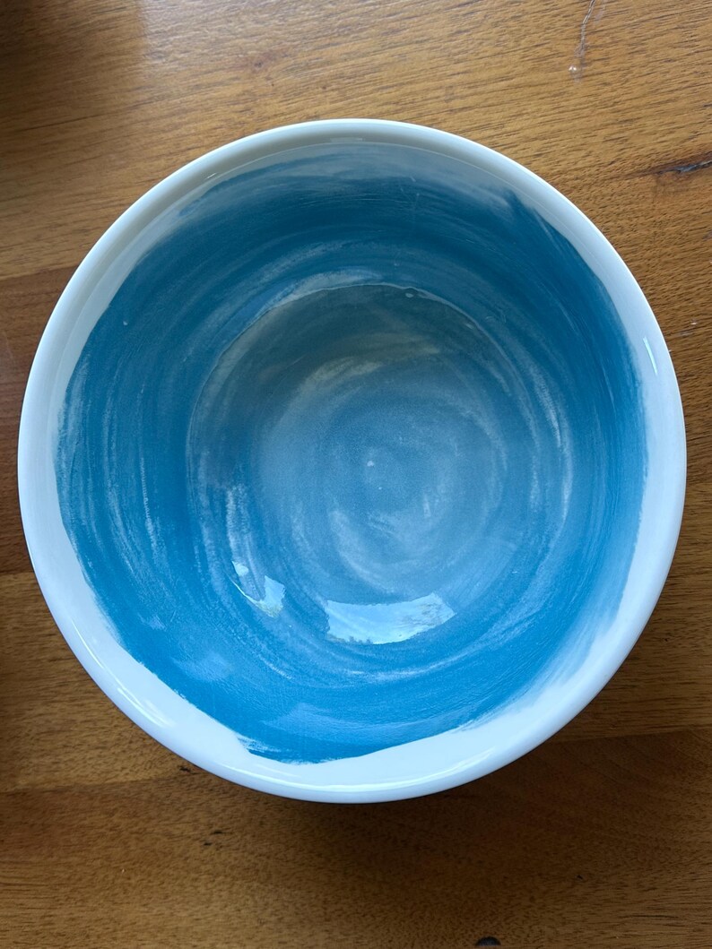 May include: A round ceramic bowl with a white rim and a blue interior. The blue paint has a brushstroke texture, creating a swirling pattern. The bowl is set against a wooden surface.