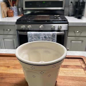 Könnte beinhalten: Cremefarbener Keramik-Blumentopf mit blauem Rand und kleinen blauen Tupfen. Der Topf hat eine einzigartige, geometrische Form und steht auf einem Holzschneidebrett in einer Küche. Der Topf ist leer.