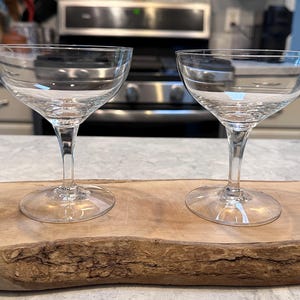 Puede incluir: Dos copas de champán transparentes con cuencos anchos y poco profundos. Las copas están sobre una tabla de madera natural. El fondo incluye una cocina con un horno de acero inoxidable.