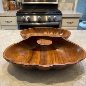 Puede incluir: Dos cuencos de madera para servir con borde festoneado y asa. Los cuencos son de madera marrón claro y tienen un patrón de vetas de madera natural. Los cuencos están sobre una encimera gris claro.