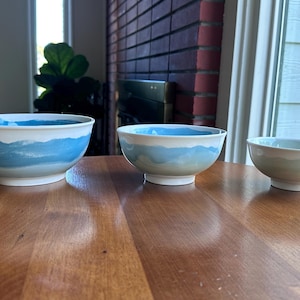 May include: Three ceramic bowls with a white base and a blue watercolor design. The bowls are arranged on a wooden table. The largest bowl is approximately 10 inches in diameter, the medium bowl is 8 inches, and the smallest is 6 inches.