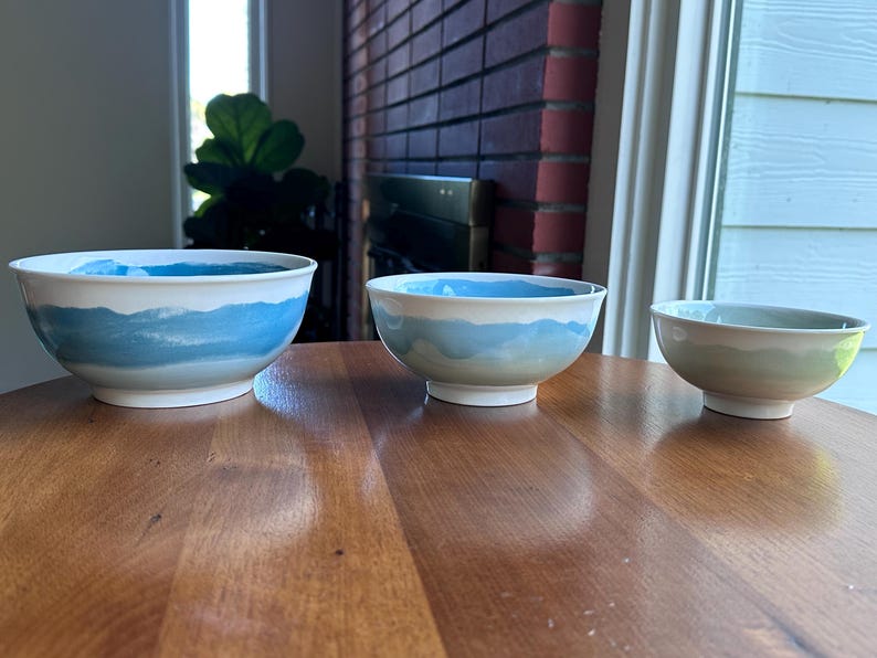 May include: Three ceramic bowls with a white base and a blue watercolor design. The bowls are arranged on a wooden table. The largest bowl is approximately 10 inches in diameter, the medium bowl is 8 inches, and the smallest is 6 inches.