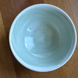 May include: A round, light green ceramic bowl with a white rim. The bowl has a smooth, glossy finish and is set against a wooden surface. The bowl's interior has a subtle, marbled effect.