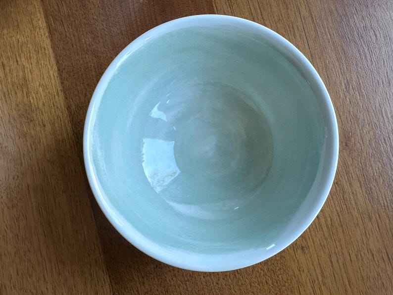 May include: A round, light green ceramic bowl with a white rim. The bowl has a smooth, glossy finish and is set against a wooden surface. The bowl's interior has a subtle, marbled effect.