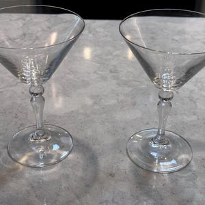 Könnte beinhalten: Zwei klare Martinigläser mit klassischem Design. Jedes Glas hat eine konische Schale, einen schlanken Stiel mit dekorativer Kugel und einen kreisförmigen Fuß. Die Gläser bestehen aus transparentem Glas.