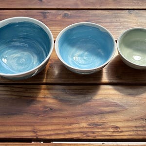 May include: Three ceramic bowls of varying sizes. The two larger bowls are painted with shades of blue, while the smallest bowl is a light green. All bowls have a white rim and sit on a wooden surface.