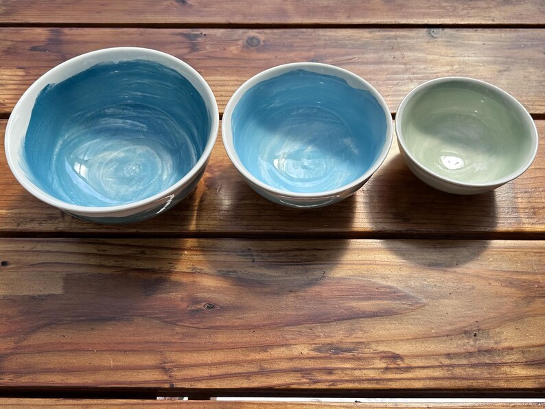 May include: Three ceramic bowls of varying sizes. The two larger bowls are painted with shades of blue, while the smallest bowl is a light green. All bowls have a white rim and sit on a wooden surface.