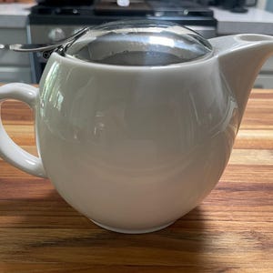 May include: A white ceramic teapot with a stainless steel infuser and lid. The teapot has a rounded body, a curved handle, and a spout. It sits on a wooden surface, suggesting a kitchen setting.