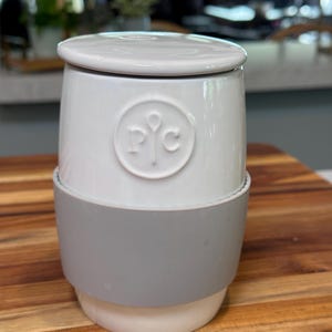 May include: A white ceramic butter dish with a matching lid. The dish features a grey silicone band and a circular logo. The butter dish is on a wooden surface.