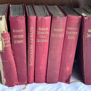 May include: A collection of seven vintage Baedeker travel guidebooks with red cloth covers and gold lettering. The titles include Southern Germany, Berlin, Süd-Bayern, Tirol, Salzburg, Austria, Eastern Alps, and Switzerland, showing signs of wear.