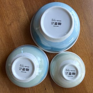 May include: Three ceramic bowls in varying sizes, each with a white interior and a blue and white watercolor-style exterior. The bowls are arranged on a wooden surface, with the bottoms of the bowls facing up, revealing the brand name and care instructions.