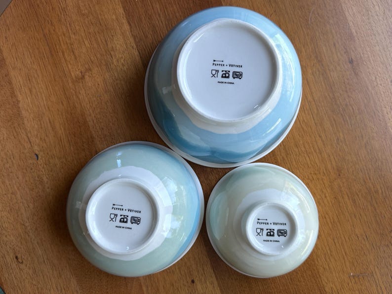 May include: Three ceramic bowls in varying sizes, each with a white interior and a blue and white watercolor-style exterior. The bowls are arranged on a wooden surface, with the bottoms of the bowls facing up, revealing the brand name and care instructions.