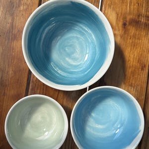 May include: Three ceramic bowls with a white exterior and a painted interior. The bowls have a blue and green color scheme. The largest bowl has a dark blue spiral design, while the other two have lighter blue and green designs.