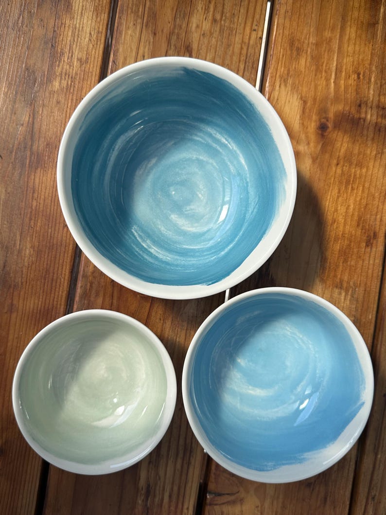May include: Three ceramic bowls with a white exterior and a painted interior. The bowls have a blue and green color scheme. The largest bowl has a dark blue spiral design, while the other two have lighter blue and green designs.