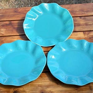 May include: Three round ceramic plates in a vibrant turquoise color. Each plate features a smooth, flat center and a distinctively ruffled, wavy edge design that creates a decorative border around the circumference of the dinnerware pieces.