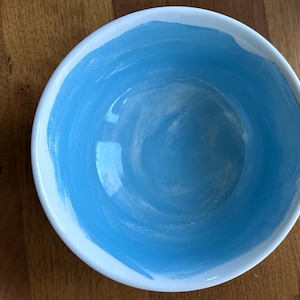 May include: A white ceramic bowl with a light blue interior. The blue paint is applied in a brushstroke pattern around the inside of the bowl. The bowl is sitting on a wooden surface.