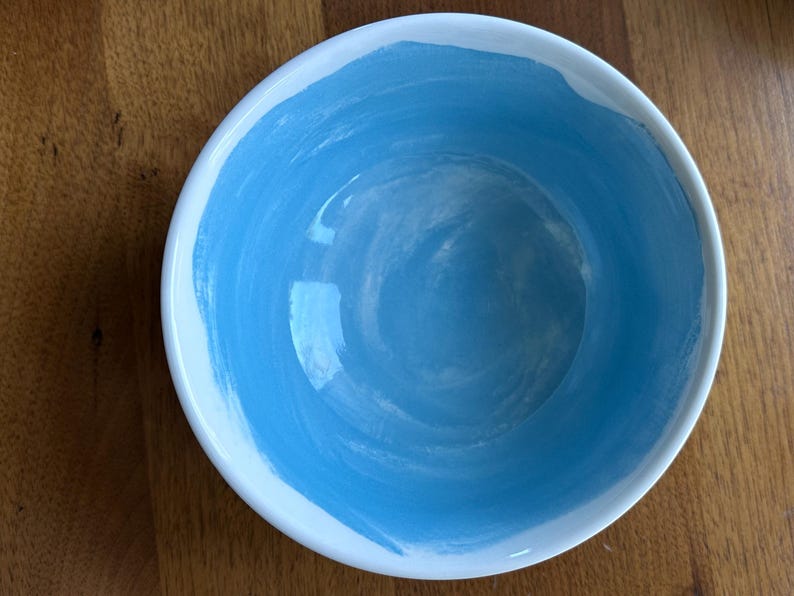 May include: A white ceramic bowl with a light blue interior. The blue paint is applied in a brushstroke pattern around the inside of the bowl. The bowl is sitting on a wooden surface.