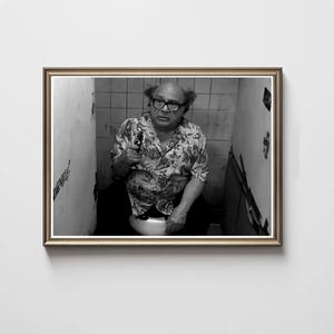 May include: Black and white framed print featuring a man in a floral shirt and glasses holding a gun. The man is seated in a bathroom stall. The image is a photograph.