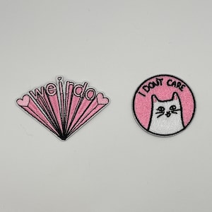 May include: Two embroidered patches. One is pink with the word "weirdo" and heart accents. The other is a pink circle with a white cat and the words "I don't care".