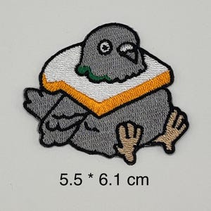May include: Embroidered patch of a grey pigeon wearing a sandwich. The patch has a white and orange border, with the text "5.5 * 6.1 cm" at the bottom. The pigeon has a green accent on its neck.