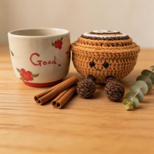 Puede incluir: Un rollo de canela tejido a ganchillo con una cara sonriente, junto a una taza de cerámica con la palabra "Good" y detalles florales. También hay palitos de canela y piezas decorativas de ganchillo marrón.
