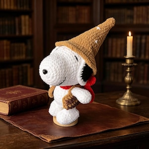 May include: A white crocheted beagle plush toy wearing a tan pointed hat with small white star accents, a red scarf, and a small brown textured satchel. The plush features black ears, a black nose, and a stitched smile, standing on a circular base.