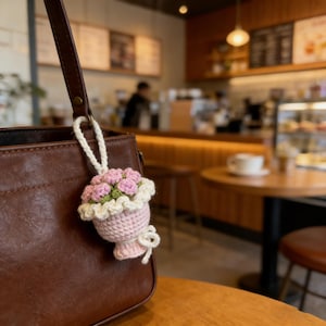 K&ouml;nnte beinhalten: Eine braune Lederhandtasche mit einem geh&auml;kelten Blumenstrau&szlig;-Schl&uuml;sselanh&auml;nger. Der Schl&uuml;sselanh&auml;nger zeigt rosa und gr&uuml;ne Blumen in einer rosa Vase mit wei&szlig;em Rand. Die Tasche h&auml;ngt an einem Holztisch in einem Caf&eacute;.